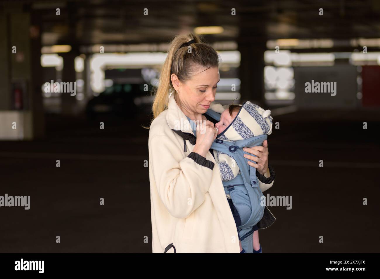 Seitenansicht einer glücklichen Frau, die ihr Baby ansieht und lächelt, während sie es in einer Babytrage auf einem Parkplatz hält und trägt Stockfoto