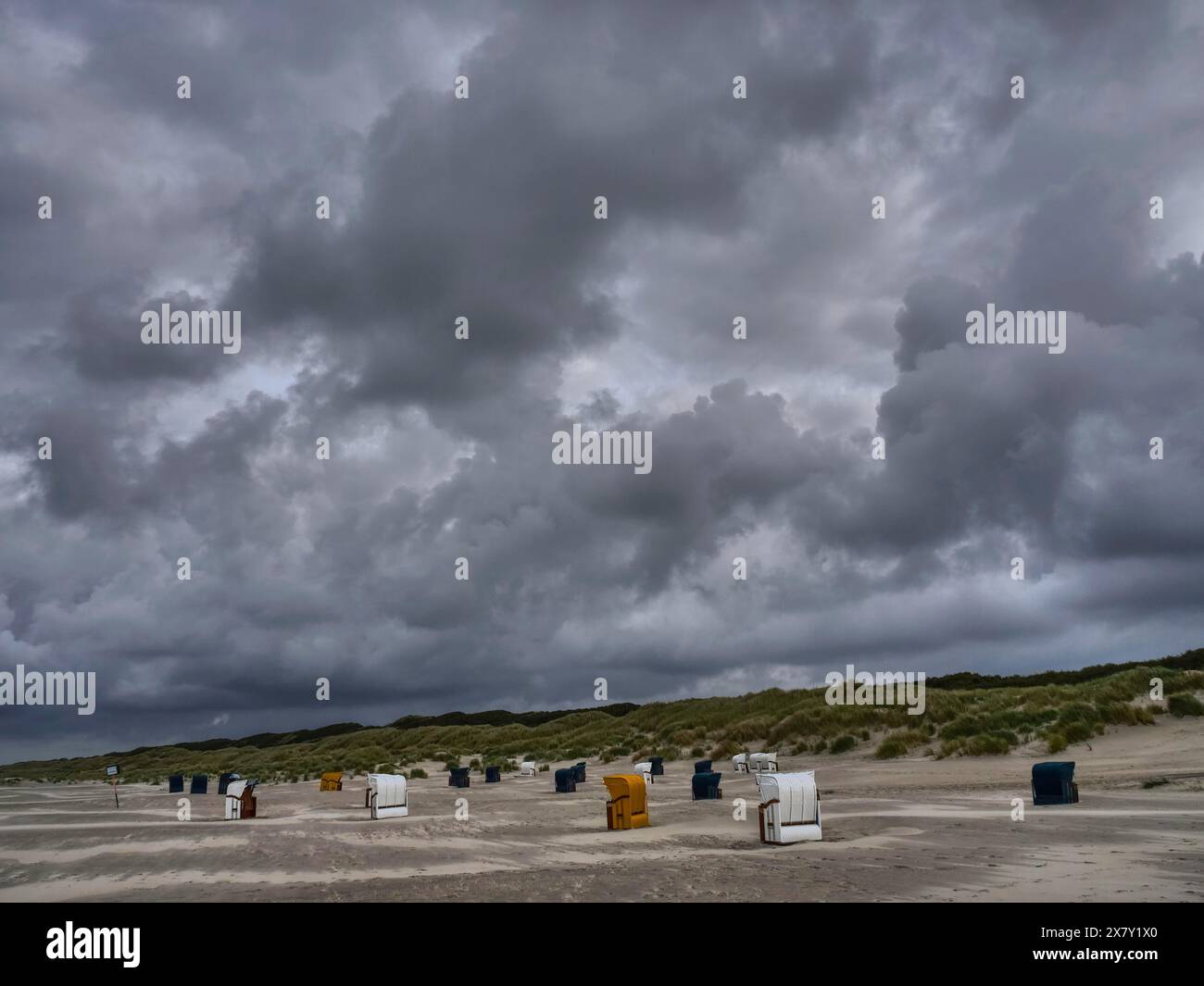 Dunkle Wolken ragen über einem grauen Strand mit verstreuten Liegen, bunten Liegen am Strand und in den Dünen unter bewölktem Himmel Stockfoto