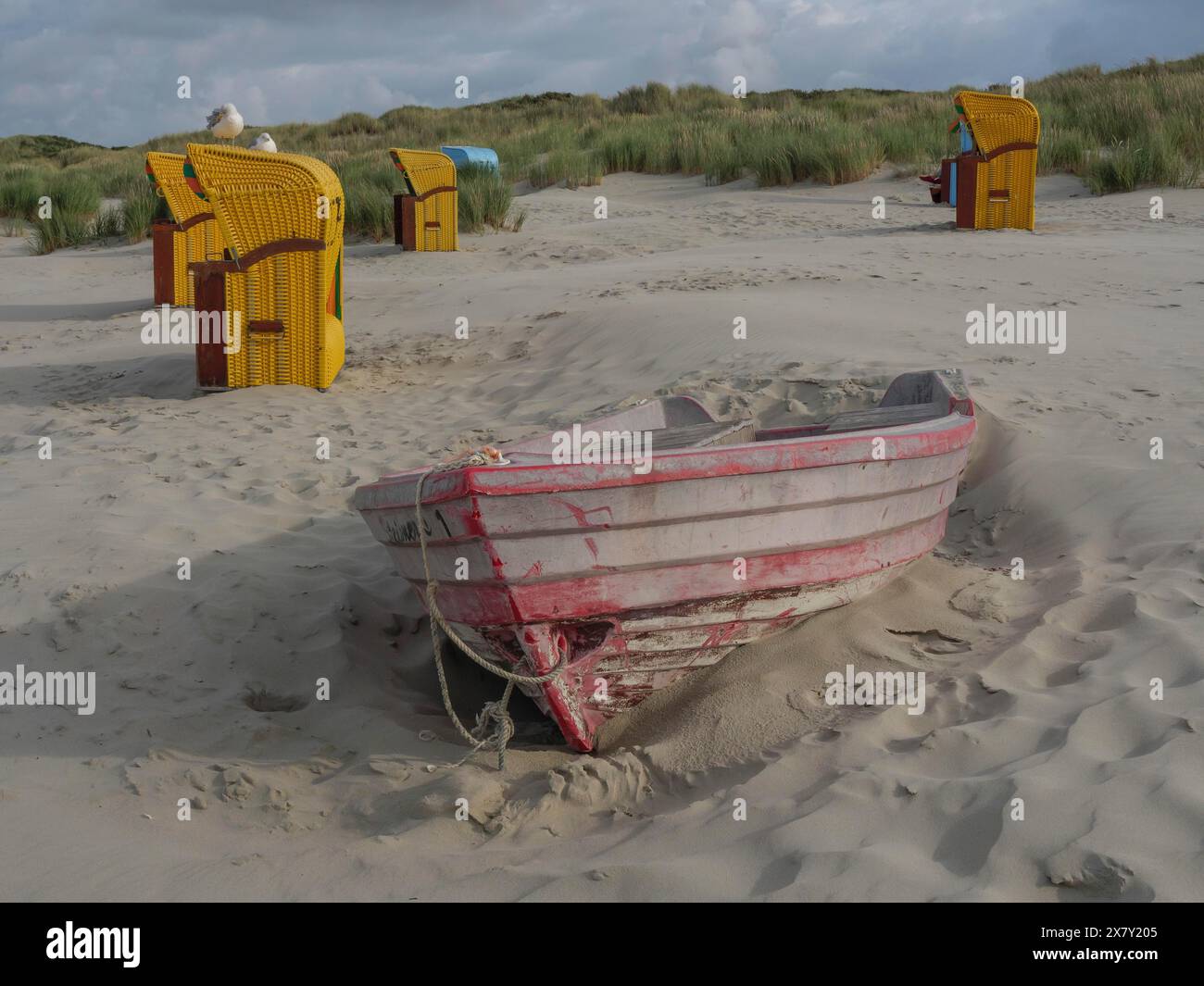 Ein verlassenes Boot, das halb im Sand versunken ist, umgeben von Liegestühlen und Dünen, bunten Liegestühlen am Strand und in den Dünen unter bewölktem Himmel, Stockfoto