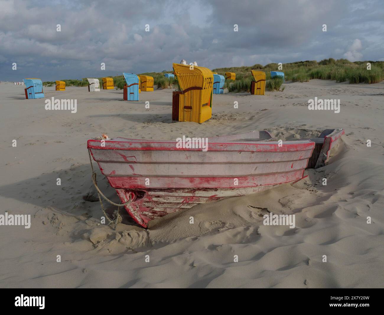 Altes, halb begrabenes Boot vor bunten Liegen am Sandstrand, bewölkter Himmel und Dünen, bunte Liegen am Strand und in den Dünen darunter Stockfoto