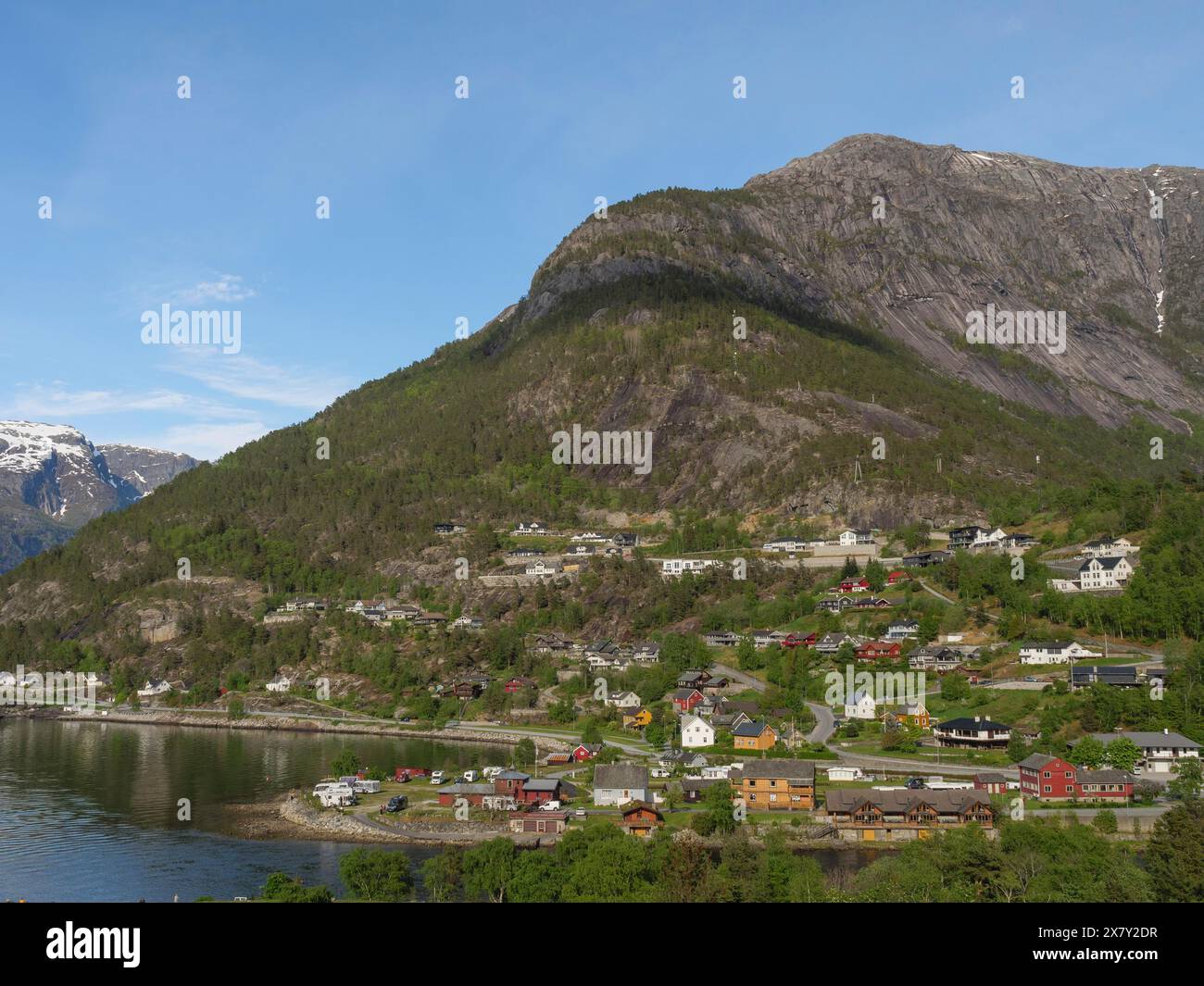 Dorf am Seeufer, eingebettet in eine Berglandschaft mit üppiger grüner Vegetation und blauem Himmel, kleines Dorf in einem Fjord vor einem hohen Berg Stockfoto