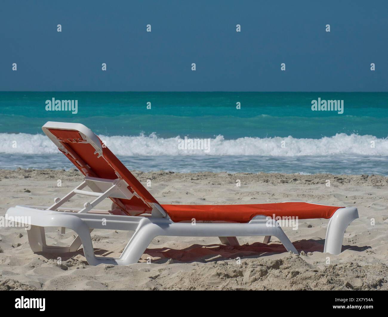 Eine einzige Sonnenliege steht schräg am Strand mit türkisfarbenem Wasser im Hintergrund und einem klaren blauen Himmel, glitzerndem grünem Meer mit Strand, Sonnen Stockfoto