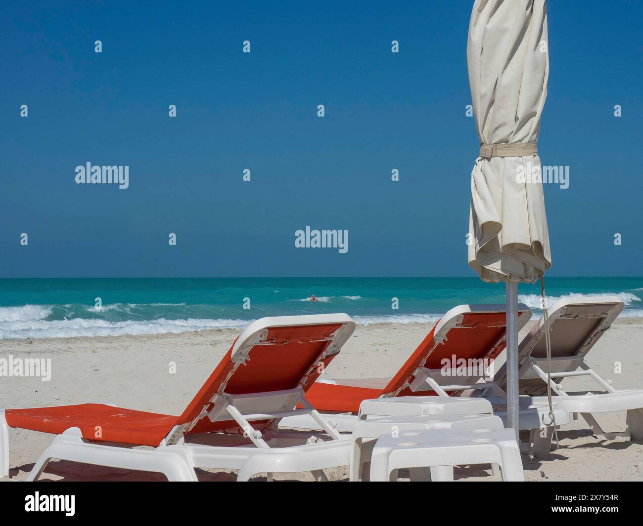Mehrere leere Liegen und ein geschlossener Sonnenschirm stehen an einem sonnigen Strand mit türkisfarbenem Wasser und blauem Himmel, glitzerndem grünem Meer mit Strand, Sonnenschirmen und Sonne Stockfoto