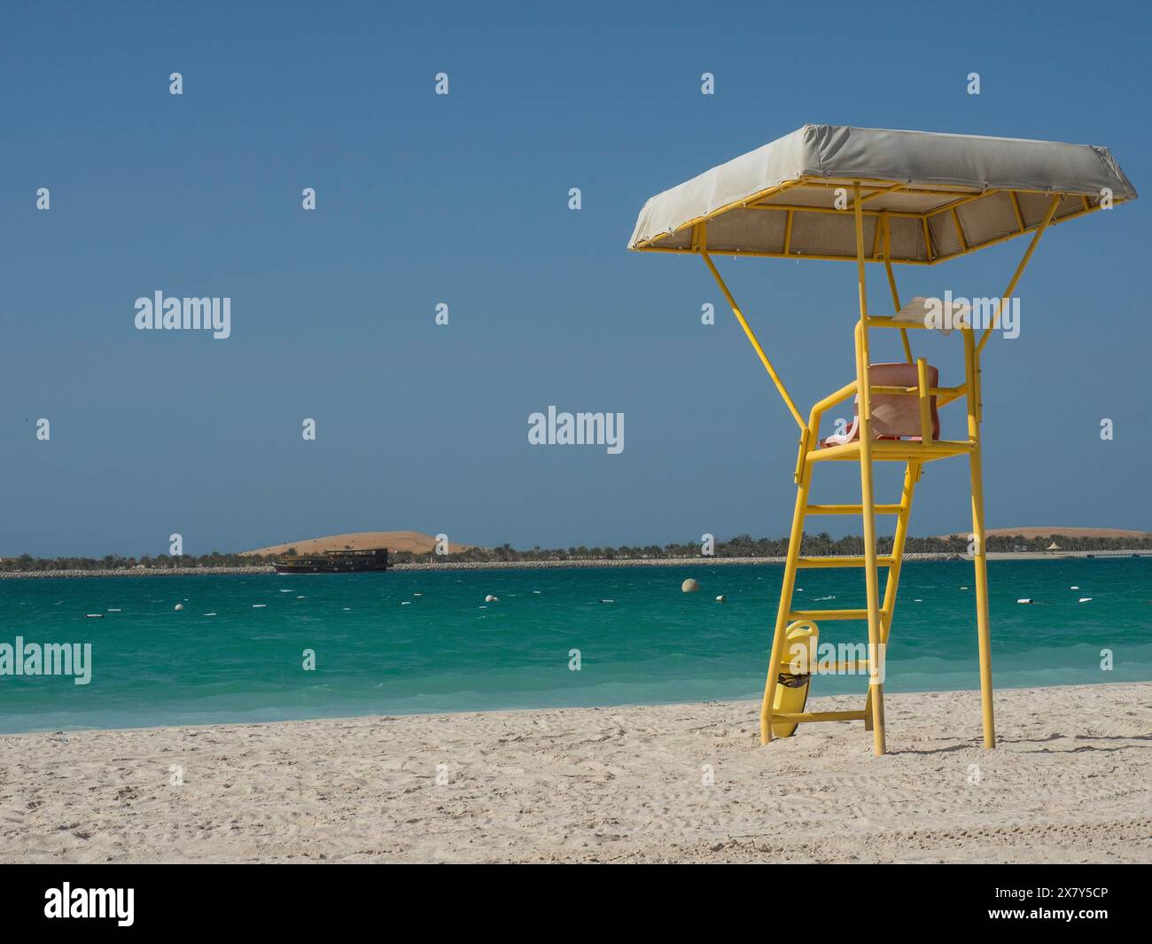 Am Strand steht ein gelber Rettungsschwimmer-Turm mit türkisfarbenem Wasser im Hintergrund und einem klaren blauen Himmel, glitzerndem grünem Meer mit Strand, Sonnenschirmen und Stockfoto