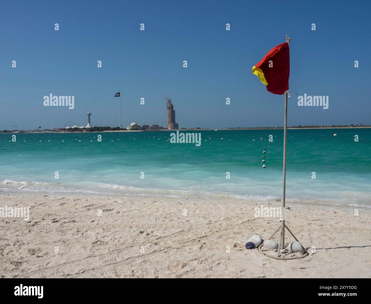 Ein rotes Warnschild steht am Strand mit türkisfarbenem Wasser und blauem Himmel im Hintergrund, glitzerndem, grünem Meer mit Strand, Sonnenschirmen und Liegestühlen Stockfoto