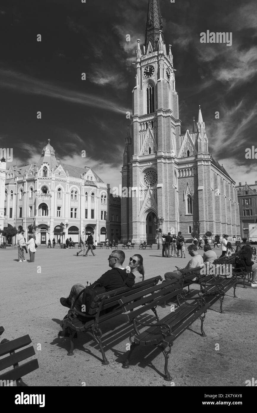 Schwarz-weiße Bänke und Menschen auf dem Freiheitsplatz, übersehen vom Turm der Kirche Name of Mary in Novi Sad, Vojvodina, Serbien Stockfoto