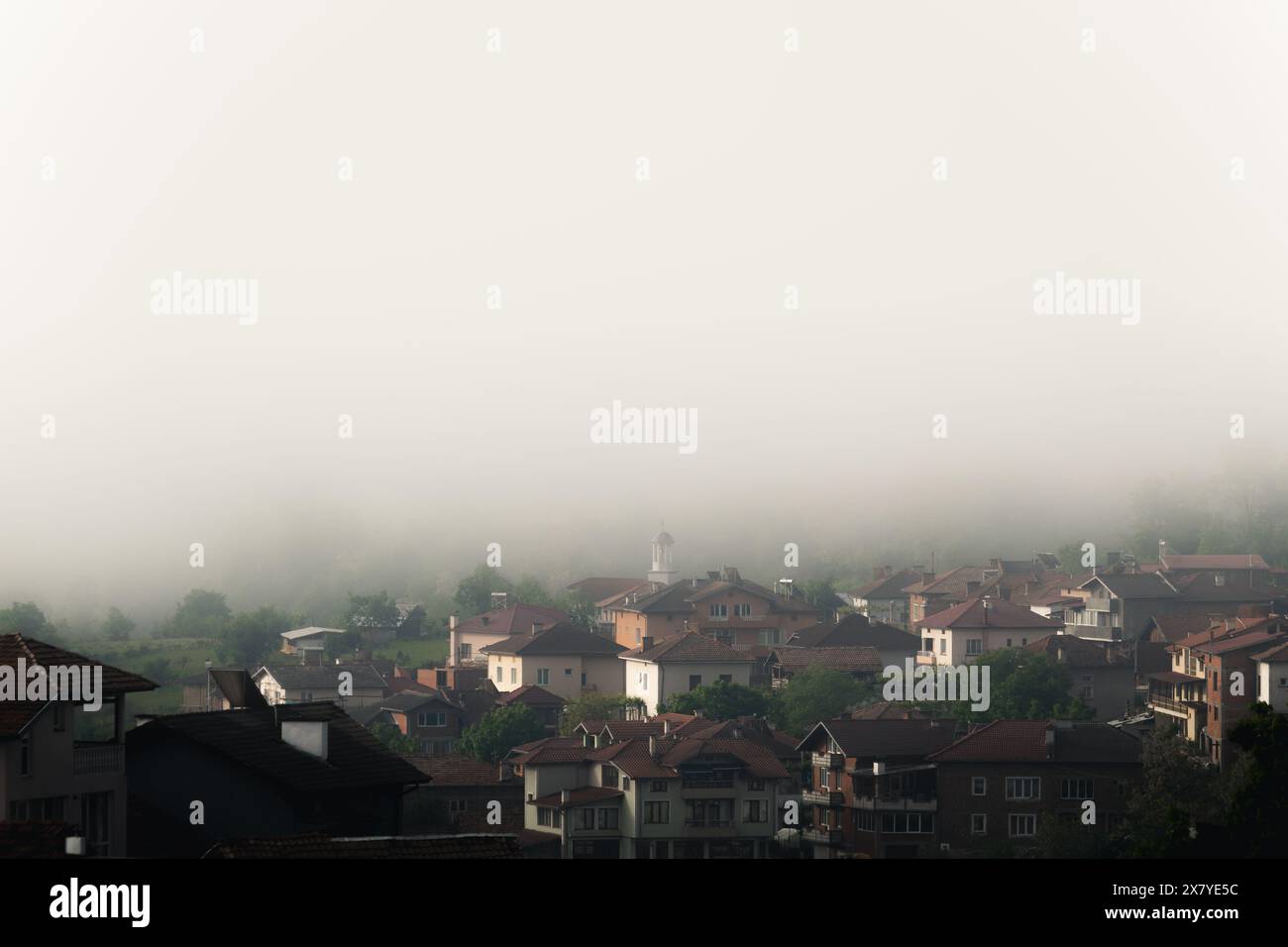 Bulgarien - 05 22 2024: Nebel über einem großen Dorf mit einer kleinen weißen Kirche in der Mitte im Hintergrund, die von Nebel bedeckt ist. Stockfoto