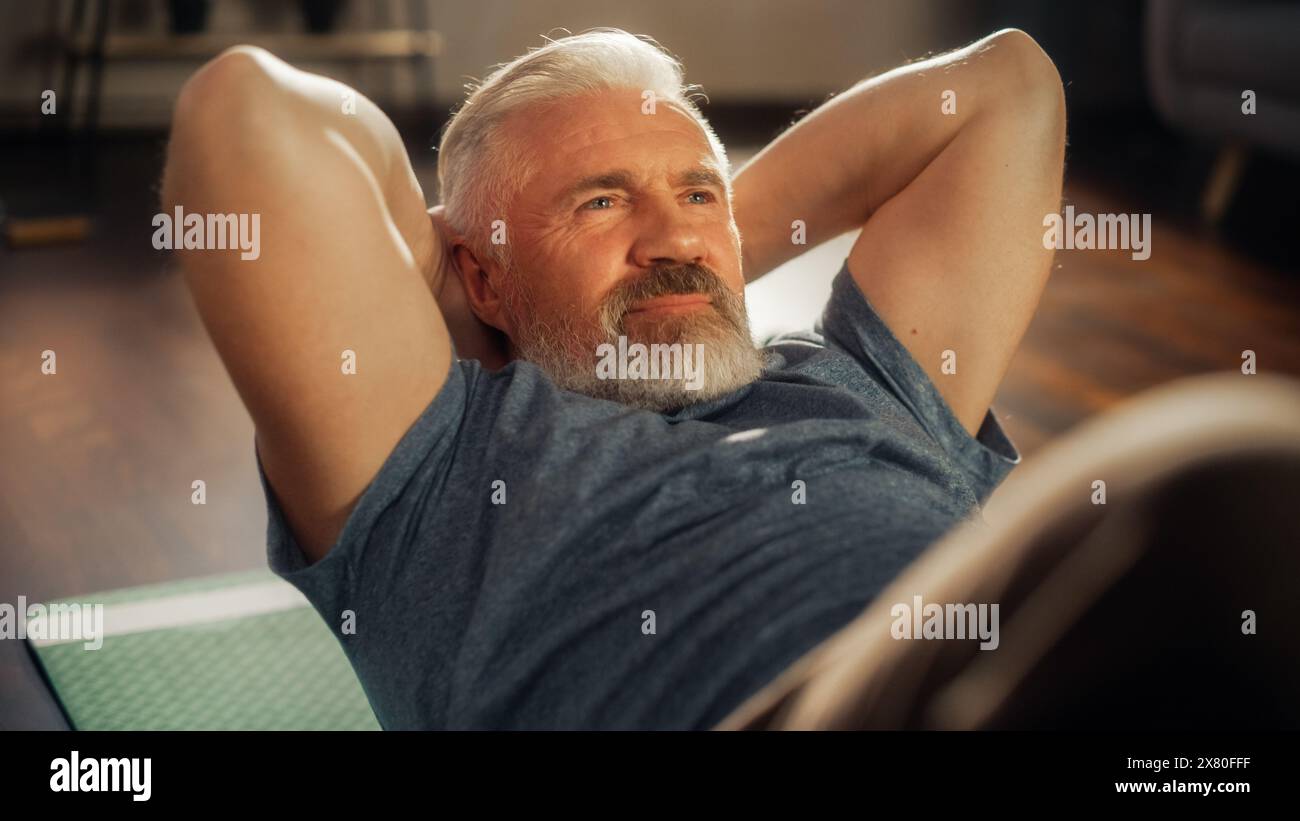 Nahaufnahme Porträt eines glücklichen Seniorenmannes, der am sonnigen Morgen zu Hause trainiert. Mann mittleren Alters, der auf dem Boden liegt und Übungen zur Stärkung des Kerns macht. Wellness und Wohlbefinden Lifestyle. Stockfoto