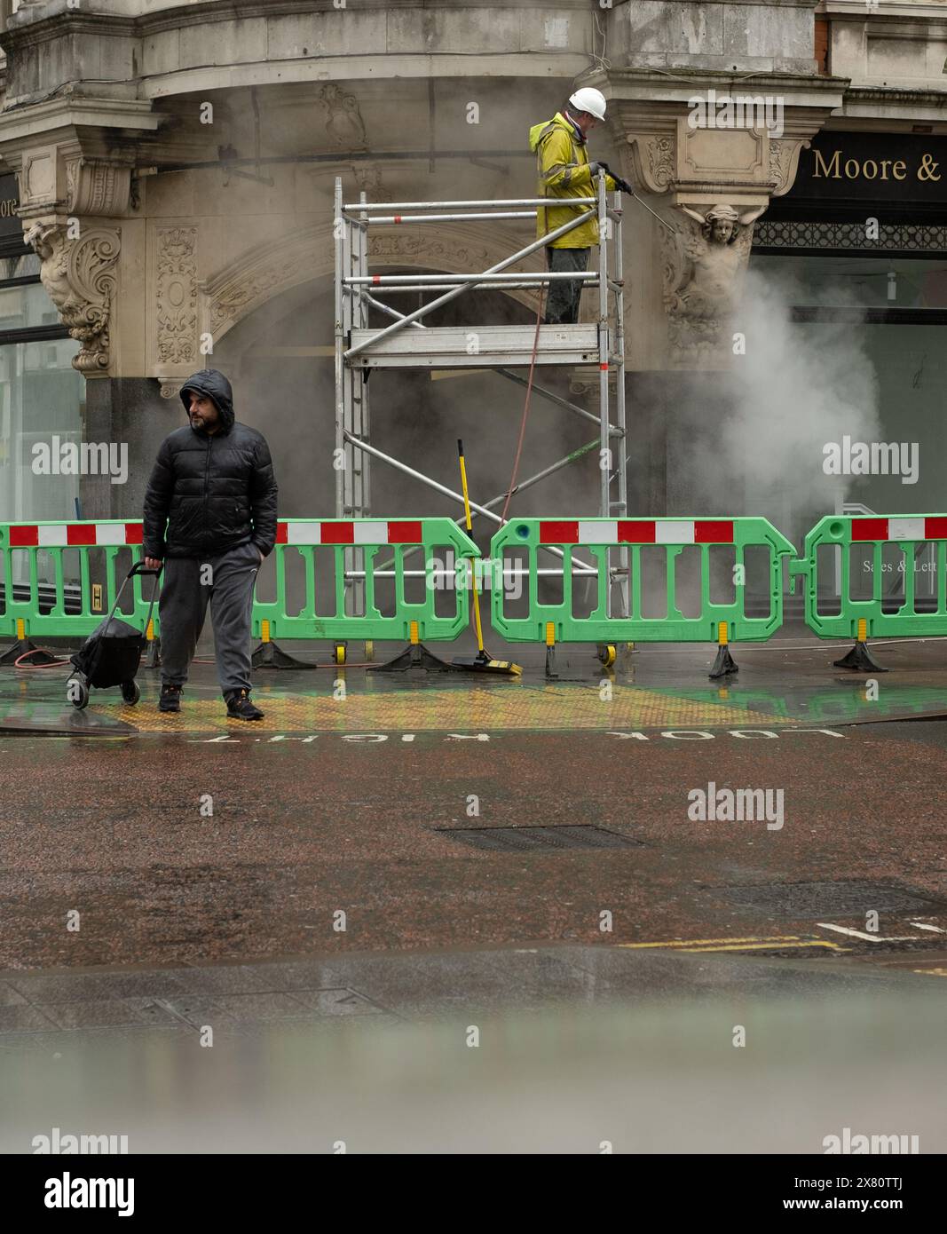 Eine Person auf der Straße in Regenkleidung, während ein Mann auf einem Gerüst das Gebäude dahinter wäscht. Straßenbarrieren und Wassernebel. Stockfoto
