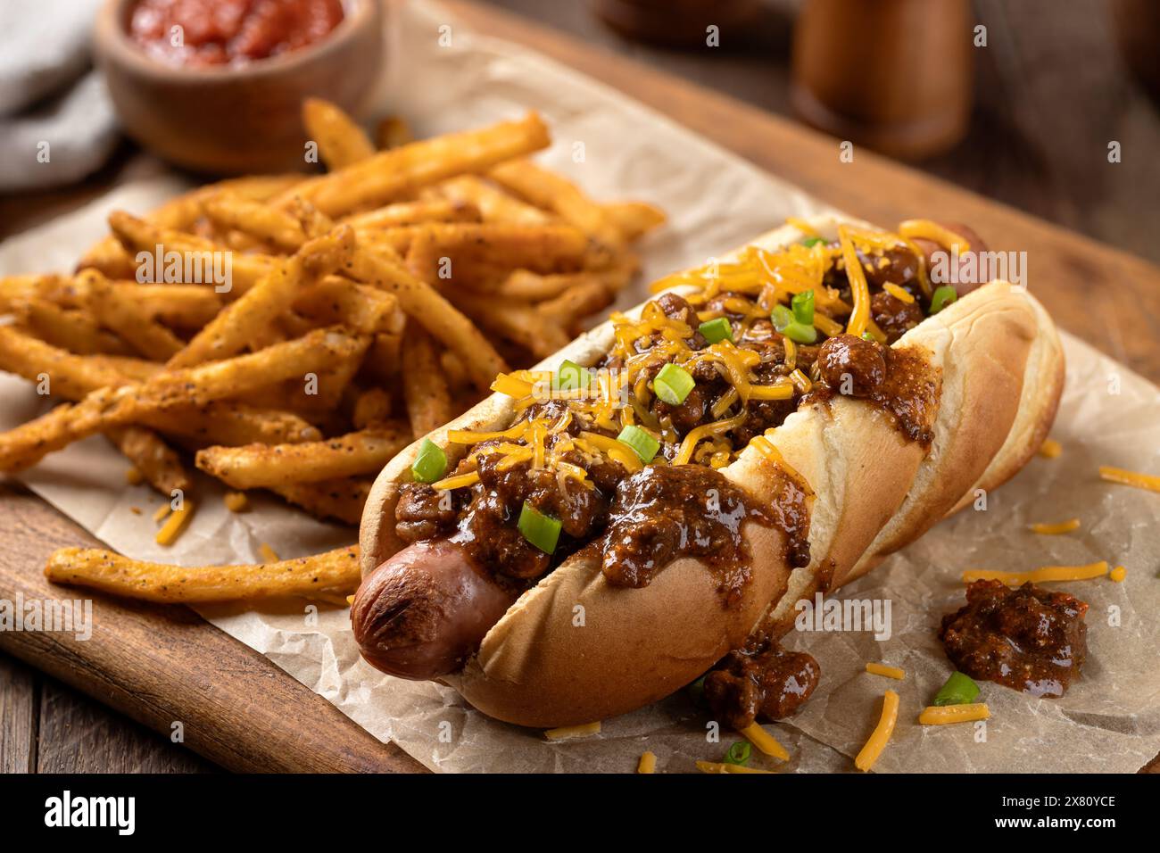 Chili Hot Dog mit geriebenem Cheddar und gewürzten Pommes frites auf einem Holzbrett Stockfoto