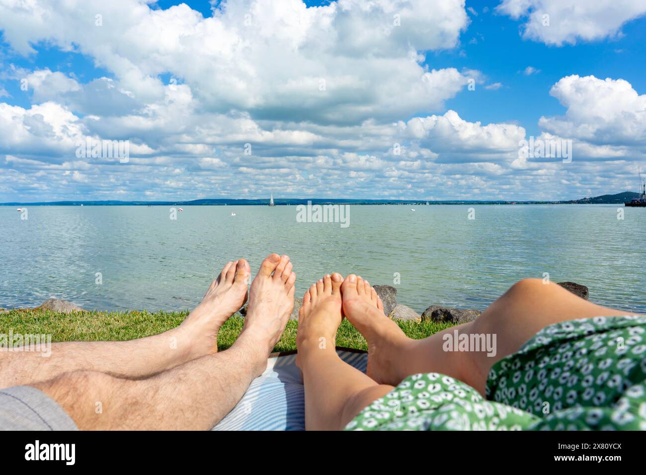 Ein Paar hängt am Strand von Badacsony mit blauem Himmel und Wolkenreflektion auf dem Wasser auf einer Decke neben dem Balaton in Ungarn ab. Stockfoto