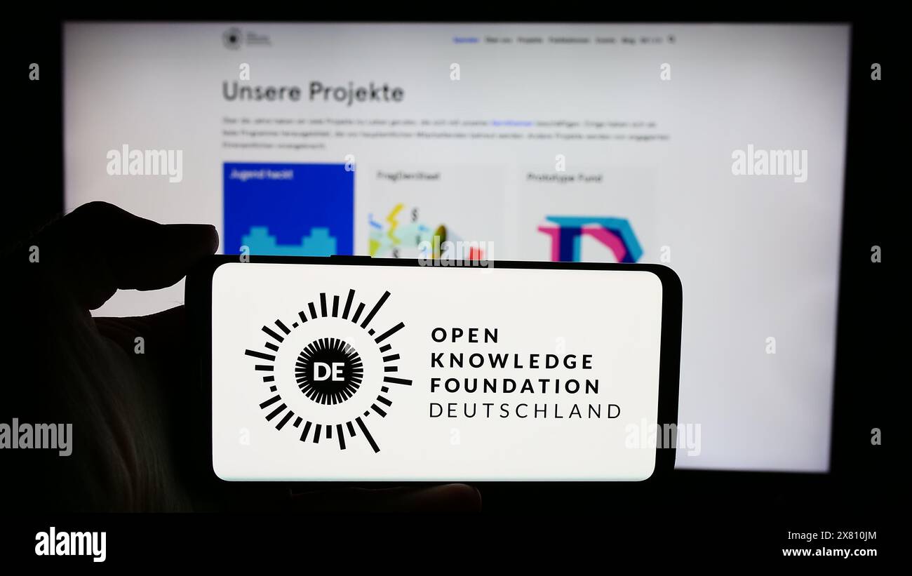 Person mit Smartphone und Logo der Open Knowledge Foundation Deutschland e.V. (OKFDE) vor der Website. Schwerpunkt auf dem Display des Telefons. Stockfoto