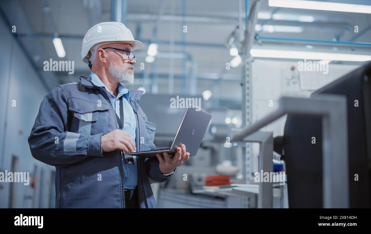 Porträt eines bärtigen General Managers mittleren Alters, der in einer Fabrik steht, einen Arbeitsanzug, eine stilvolle blaue Brille und einen weißen Schutzhelm trägt. Spezialist für Schwerindustrie, der an Laptops arbeitet. Stockfoto