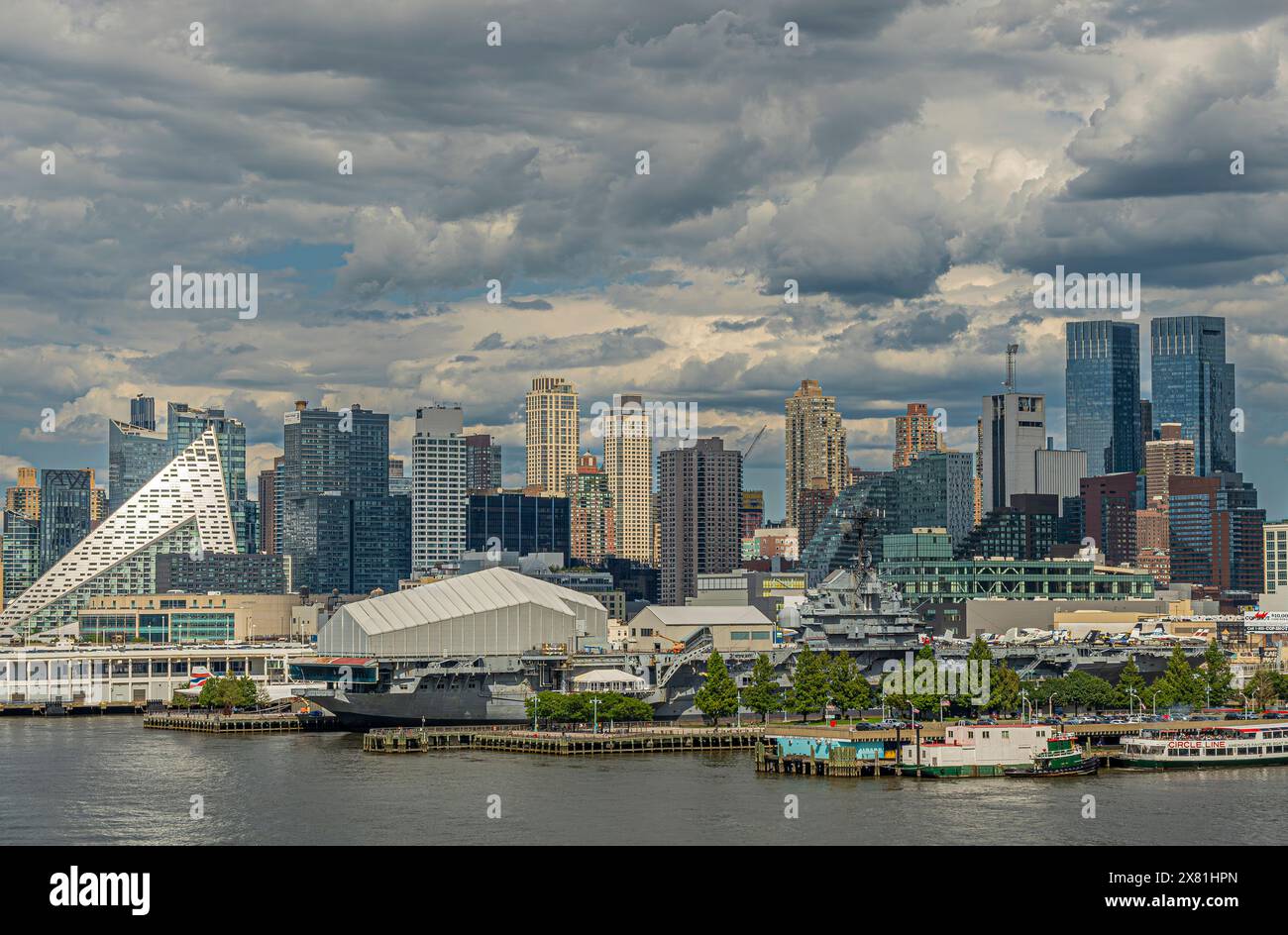 New York, NY, USA - 1. August 2023: Unerschrockener Flugzeugträger am Pier 86 mit Wolkenkratzern und anderen Gebäuden dahinter unter blaugrauer Wolkendecke Stockfoto