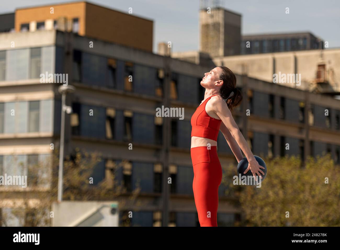 Sportliche Frau, die mit einem kleinen Pilates-Ball hinter sich steht und draußen Dehnungsübungen macht, im urbanen Hintergrund. Stockfoto