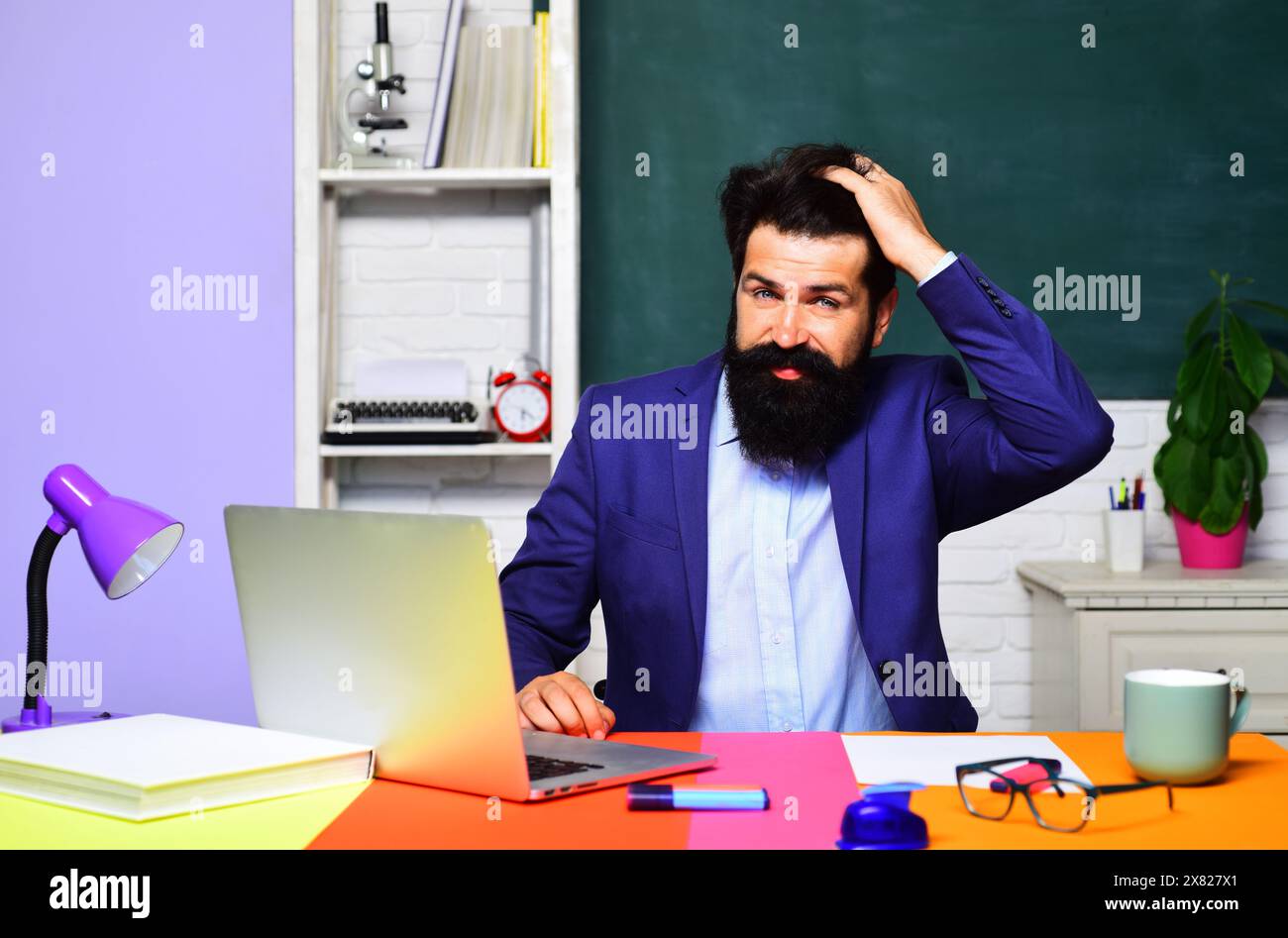 Lehrerjob. Lächelnder männlicher Lehrer im Klassenzimmer. Schöner Student auf dem Universitätscampus. Zurück zur Schule. Ein bärtiger Professor sitzt am Schreibtisch und arbeitet mit Stockfoto