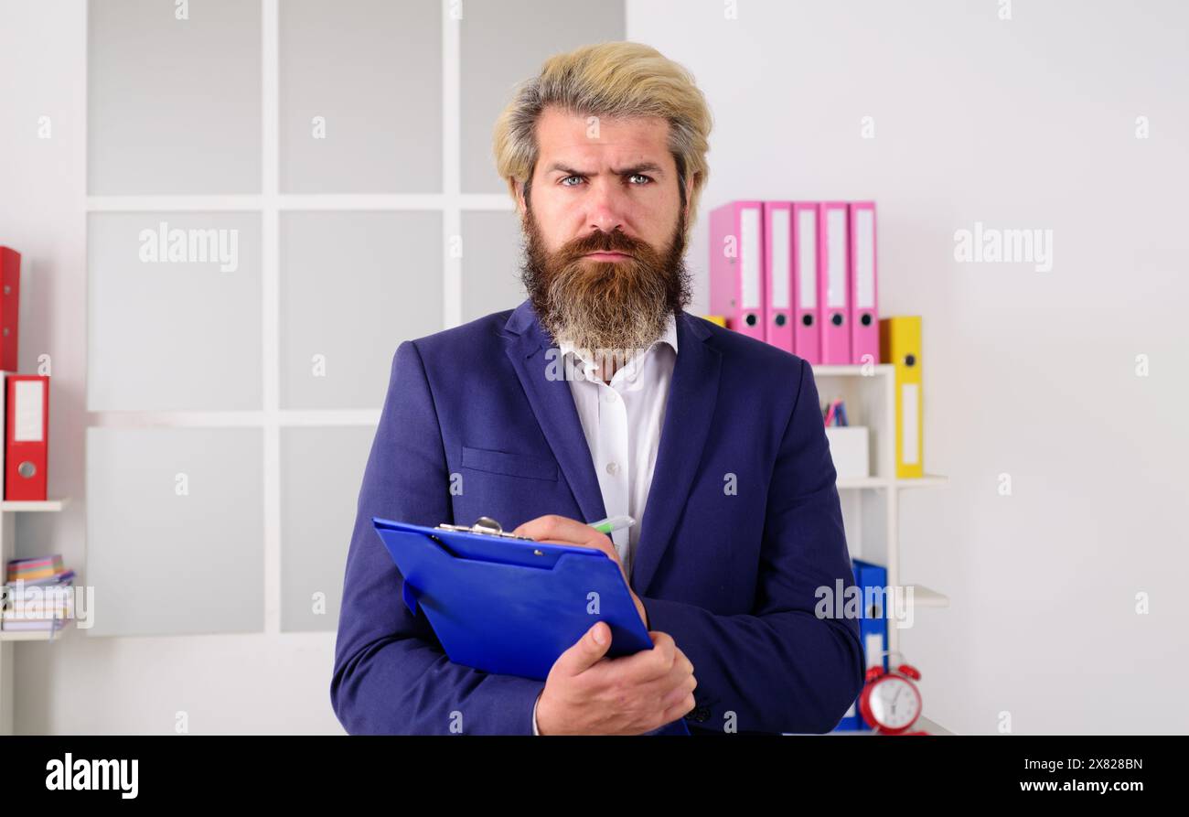 Selbstbewusster Geschäftsmann in Anzug arbeitet im Büro, hält die Zwischenablage mit Papierdokumenten, schreibt Notizen. Ernsthafter bärtiger Geschäftsmann, Unternehmensleiter Stockfoto