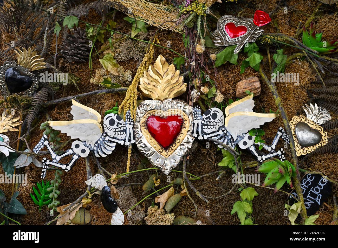 Eine Vielzahl von dekorativen Elementen, darunter kleine Schädel, ein zentrales rotes Herz und Blätter, sind in einem symmetrischen Muster gegen einen strukturierten Rücken angeordnet Stockfoto