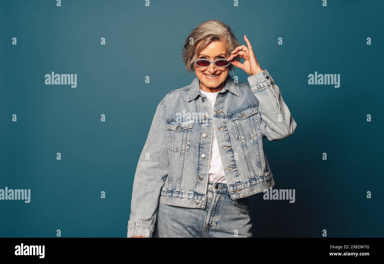 Die modische Seniorin mit grauem Haar und Brille steht in einem lässigen Denim-Outfit verspielt in einem Studio. Stockfoto