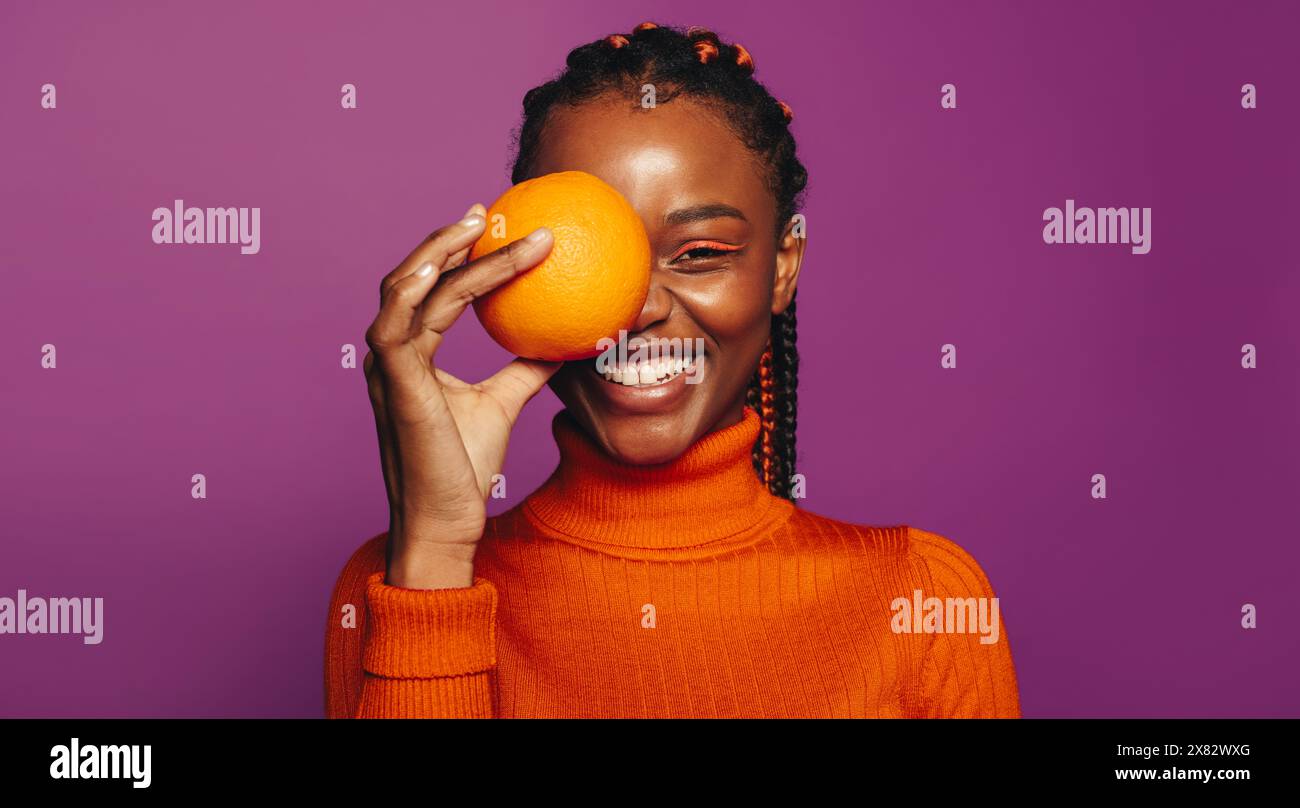 Lächelnde Afrikanerin mit verspieltem und lebendigem Stil steht in einem lila Studio. Sie hält eine Orangenfrucht, die einen gesunden Lebensstil zeigt Stockfoto