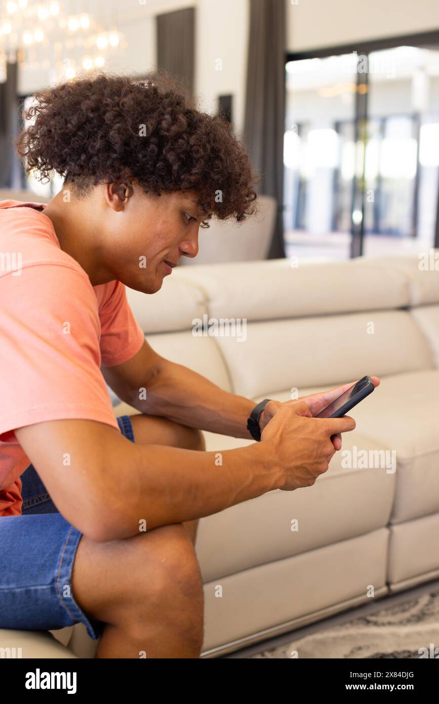 Ein birassischer junger Mann zu Hause, der auf der Couch sitzt und durch sein Smartphone blättert Stockfoto