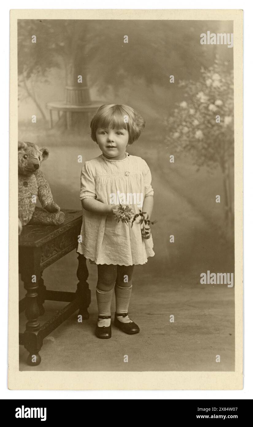 Originale charmante Studio-Postkarte aus der Zeit der 1920er oder frühen 1930er Jahre mit einem niedlichen jungen Mädchen (möglicherweise Brown-Familie), das eine Blume hält, altmodischer Teddybär auf Stuhl, gemalter Hintergrund, Kind aus den 1920er Jahren. Das Mädchen hat ein modisches, gekräuseltes Haar, trägt ein Sommerkleid. Fotostudio von H. C. Clarkson, Birkenhead, Liverpool, England, Großbritannien Stockfoto