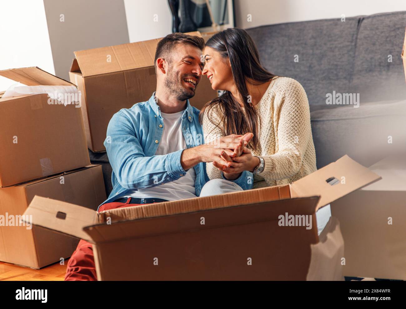 Ein lächelndes junges Paar zieht in ein neues Zuhause, sitzt auf dem Boden und packt aus. Stockfoto