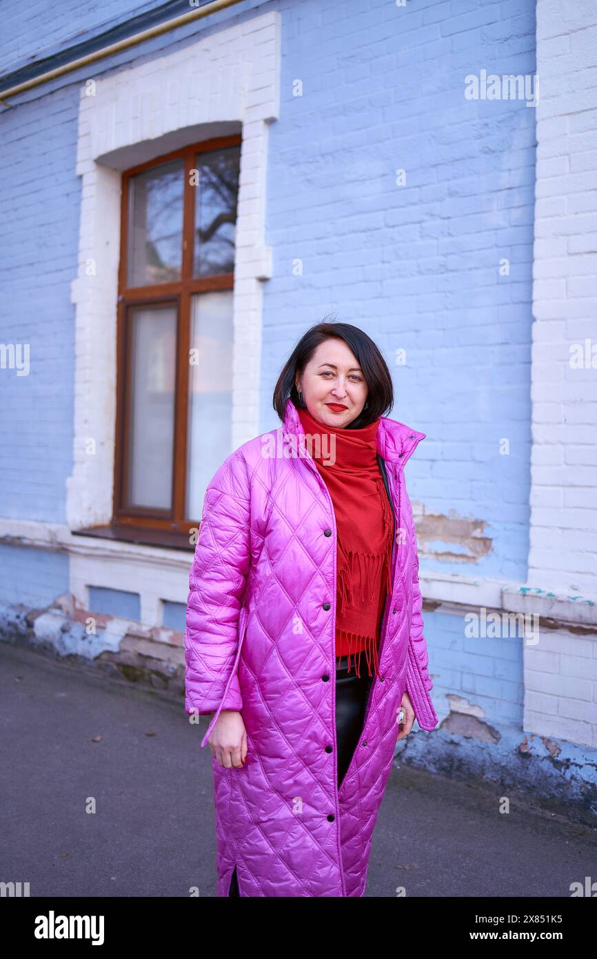Stilvolle Frau mittleren Alters in einem langen, knalligen rosa Mantel und roten Schuhen spaziert durch die Straßen der Stadt Stockfoto