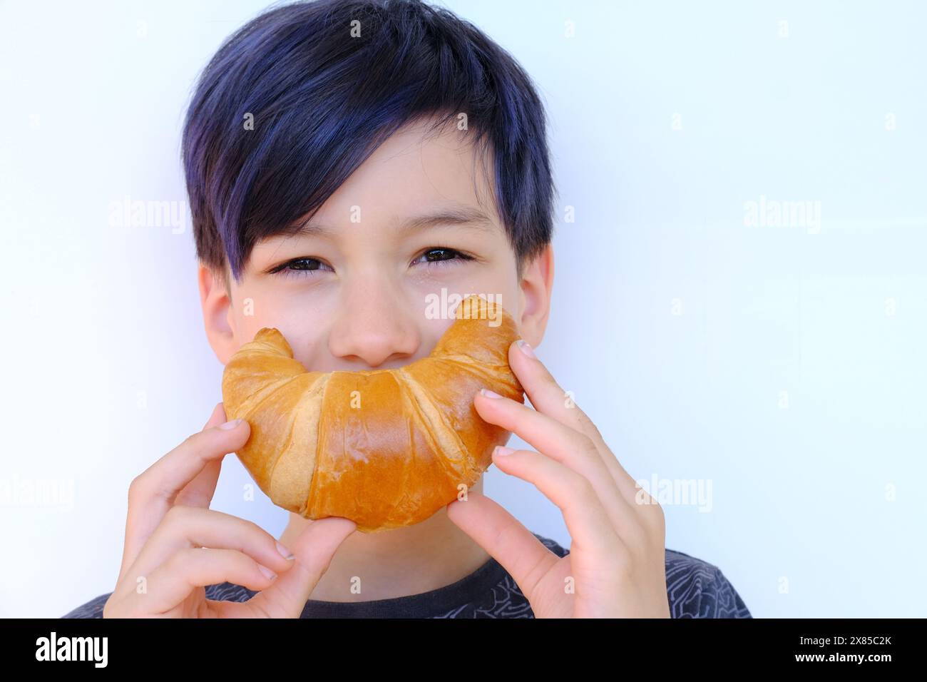 Nahaufnahme des Gesichts des Kindes, Junge von 10 Jahren hält rosa gebackenes Croissant mit Händen nahe dem Mund, zeigt fröhliches Lächeln, Konzept köstliches Frühstück, h Stockfoto