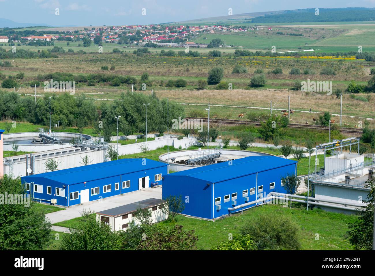 Eine Industrieanlage mit blauen Strukturen und Tanks in ländlicher Landschaft und sanften grünen Hügeln mit einem entfernten Dorf. Stockfoto