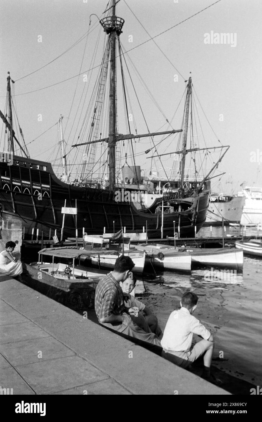 Drei junge Männer sitzen auf Stufen an der Wasserlinie im Alten Hafen von Barcelona, im Hintergrund ankert eine Galeere, Spanien 1957. Drei junge Männer sitzen auf Stufen an der Wasserlinie im Alten Hafen Barcelonas, eine Galeere im Hintergrund, Spanien 1957. Stockfoto