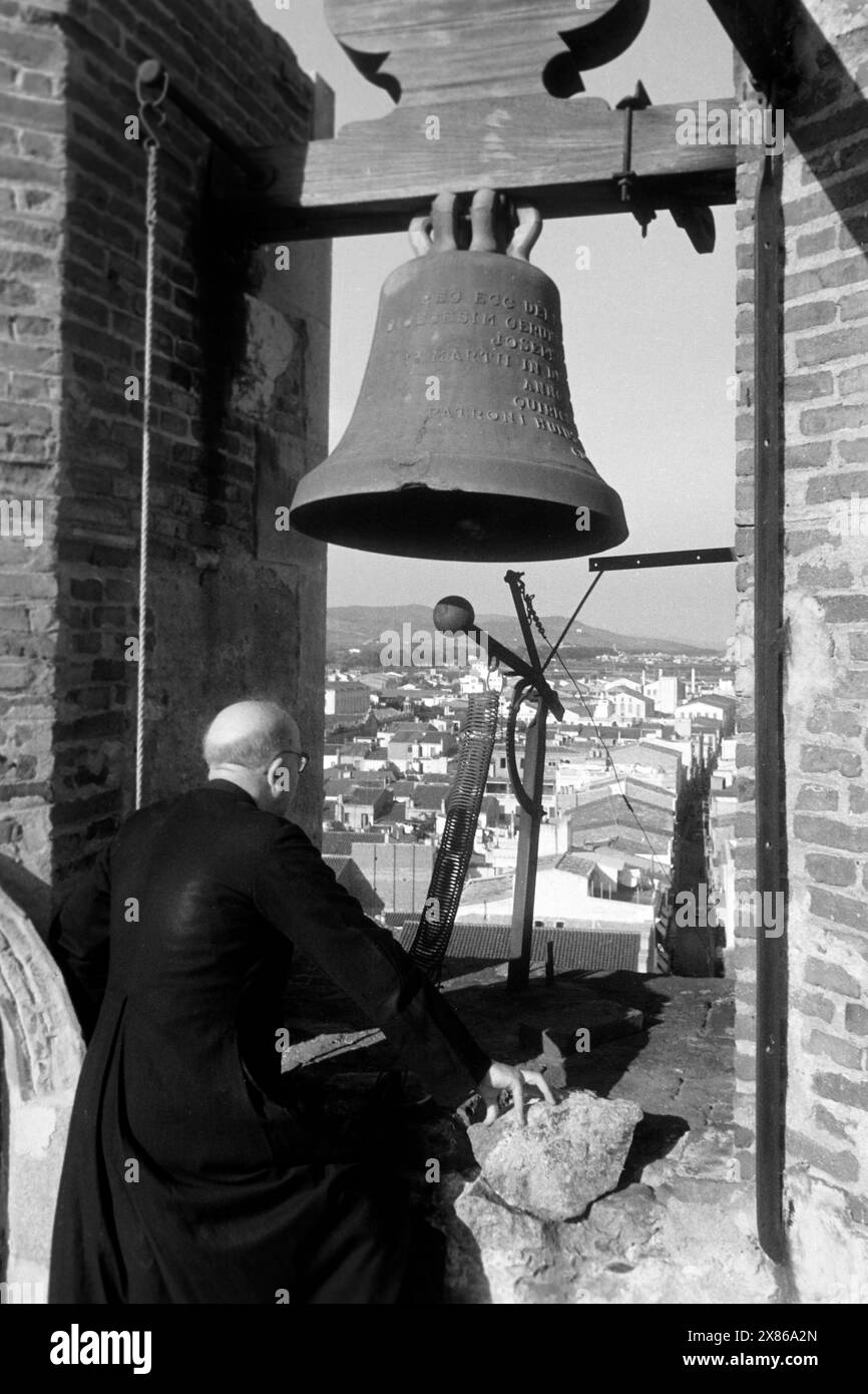 Ein Geistlicher blickt an der Glocke eines Glockenturms vorbei auf die umliegende Ortschaft von Calella an der Costa del Maresme, Katalonien 1957.Ein Geistlicher blickt an der Glocke eines Glockenturms vorbei an der umliegenden Ortschaft Calella an der Costa del Maresme, Katalonien 1957. Stockfoto