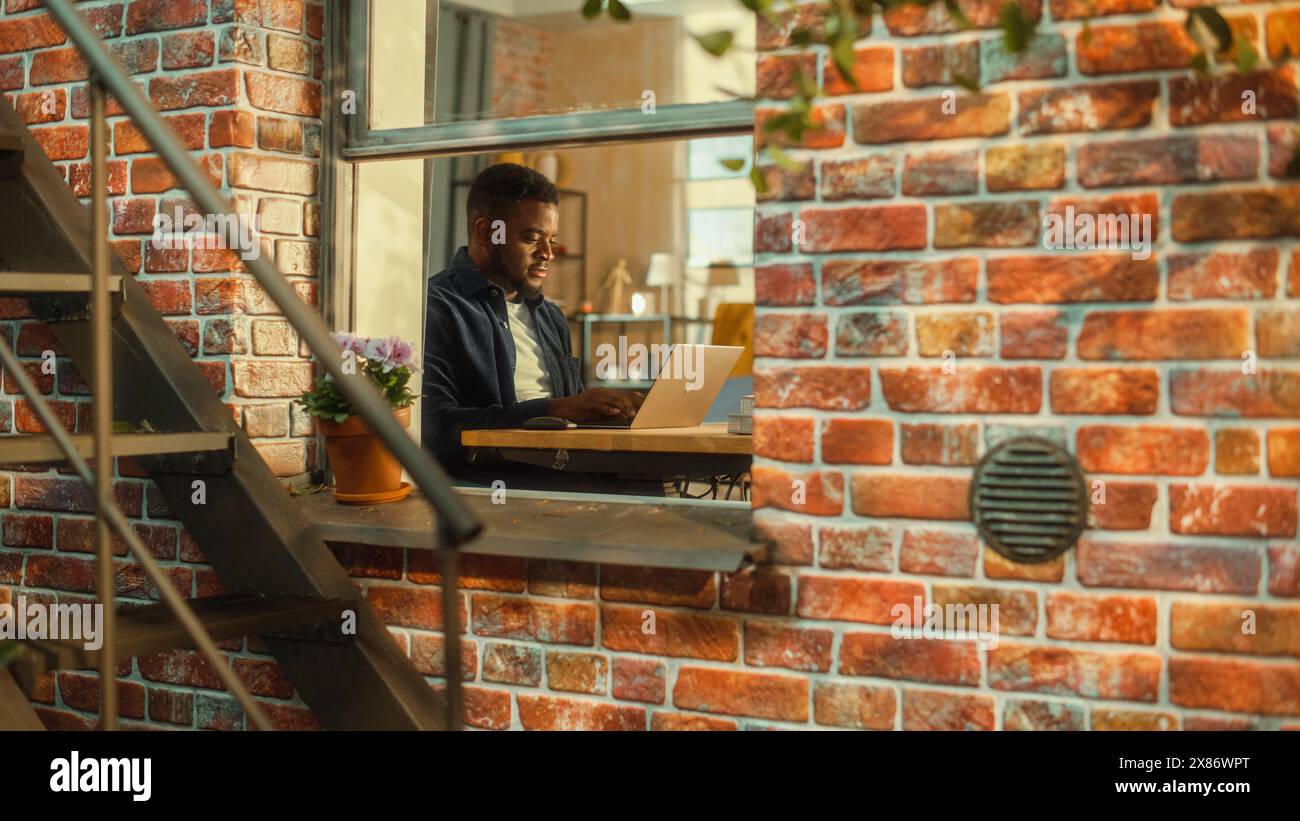 Junger Schwarzer, der zu Hause ein Notebook benutzt. Inspirierende Fernarbeit während des Tages. Spezialist, der am Computer arbeitet, einen kreativen Projektplan schreibt. Von außerhalb des Fensters geschossen. Stockfoto