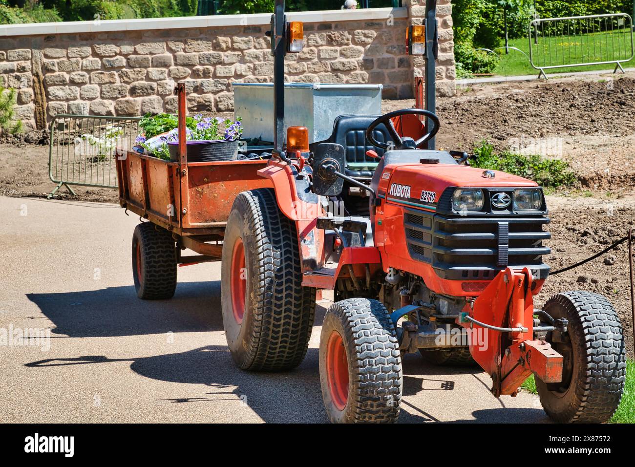 Ein roter Traktor mit einem Anhänger mit Topfpflanzen, der auf einem gepflasterten Weg in einem Gartenbereich abgestellt ist. Stockfoto