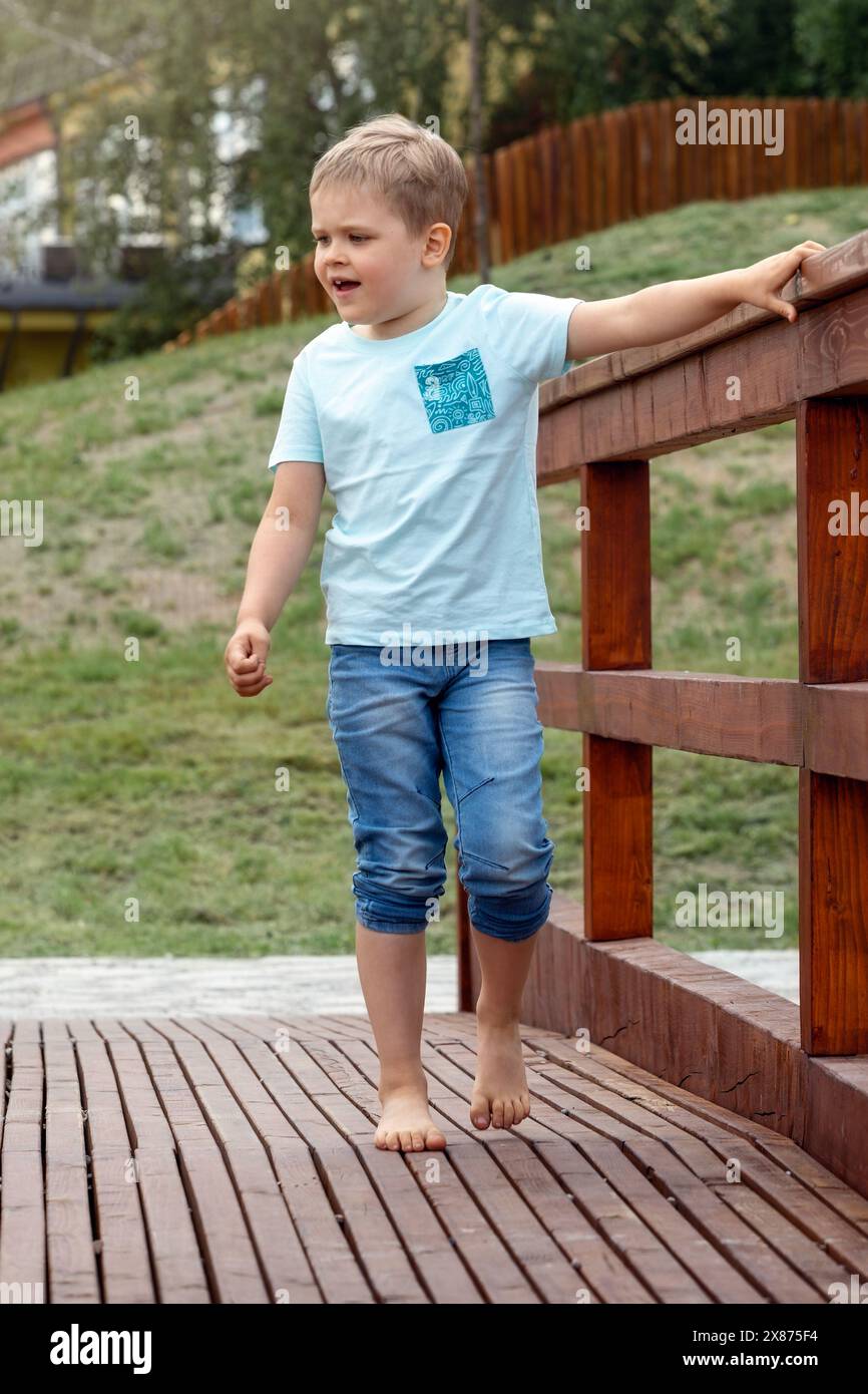 Ein kleiner Barfuß-Junge in blauer Kleidung läuft auf einer Holzbrücke in einem Stadtpark und hält sich am Geländer. Stockfoto