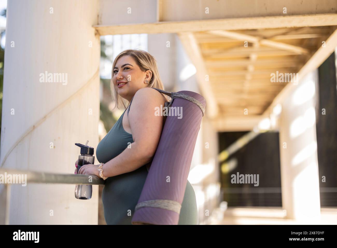 Lächelnde junge kurvige Frau, die mit Trainingsmatte und Wasserflasche steht Stockfoto