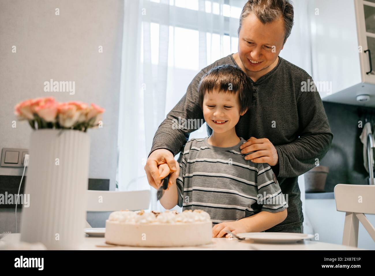 Ein lächelnder Mann schneidet Geburtstagskuchen mit seinem Sohn in der Küche zu Hause Stockfoto
