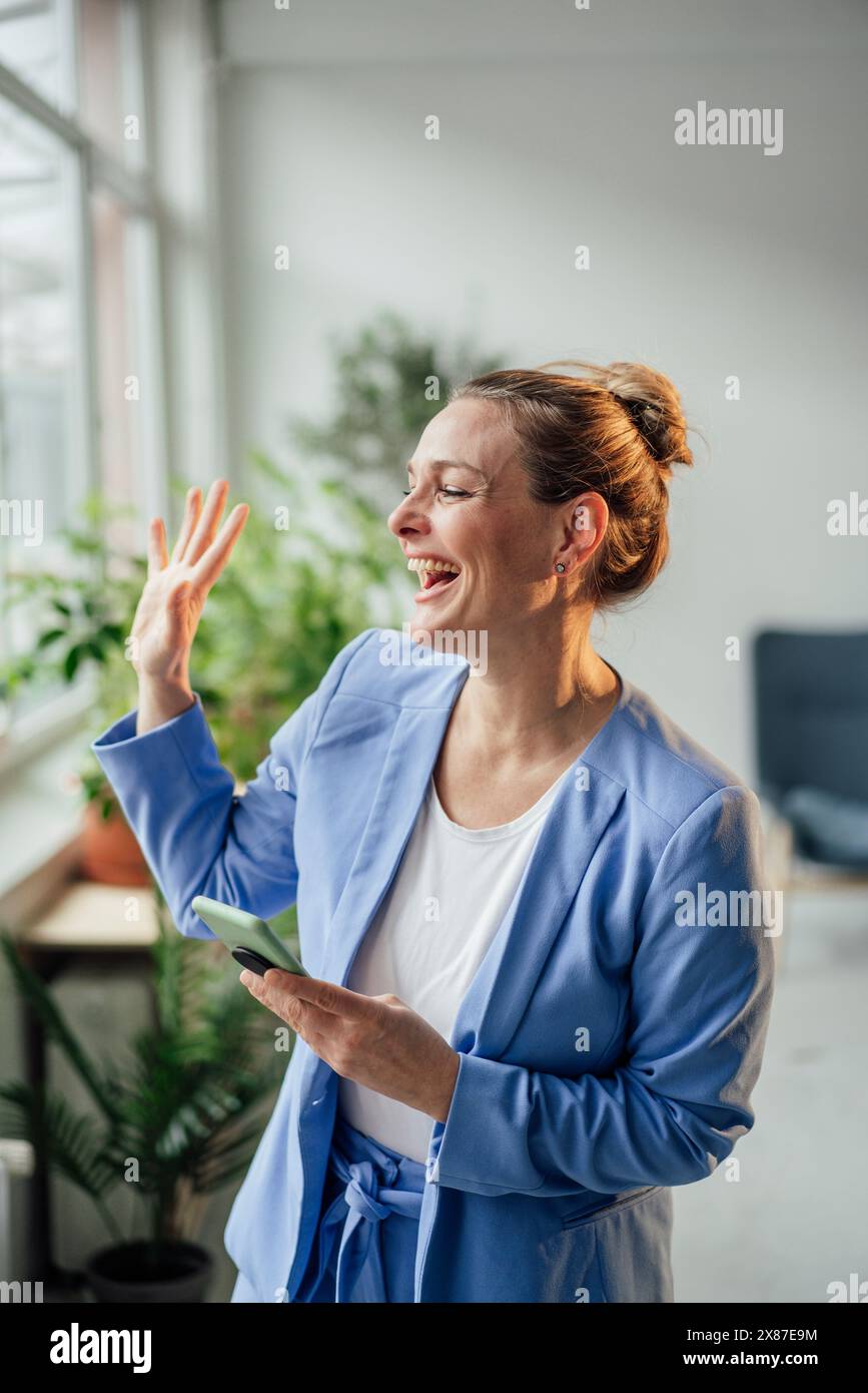 Fröhliche Geschäftsfrau mit Smartphone, das im Büro winkt Stockfoto