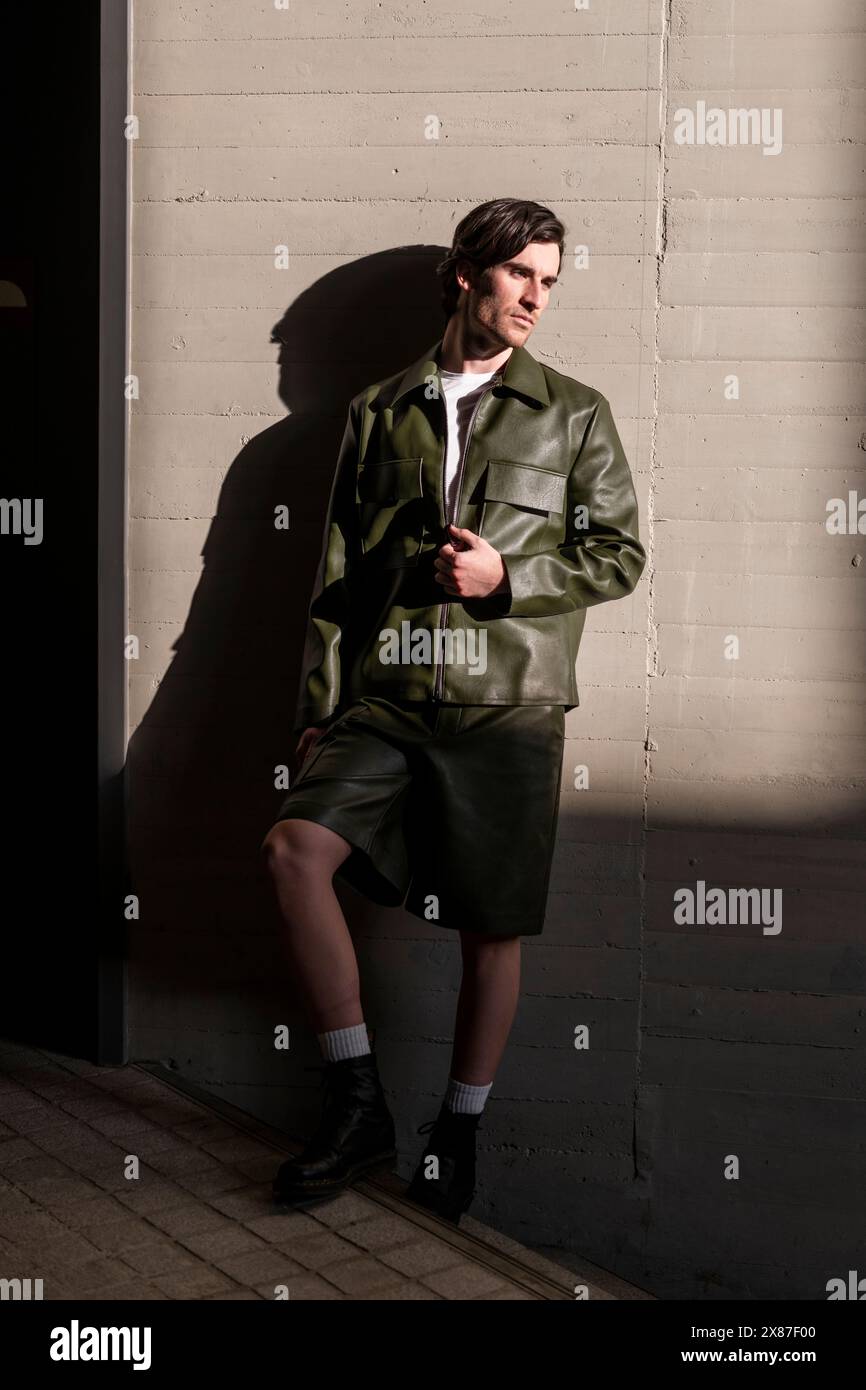 Modischer junger, schöner Mann mit grüner Lederjacke und Shorts an der Wand Stockfoto