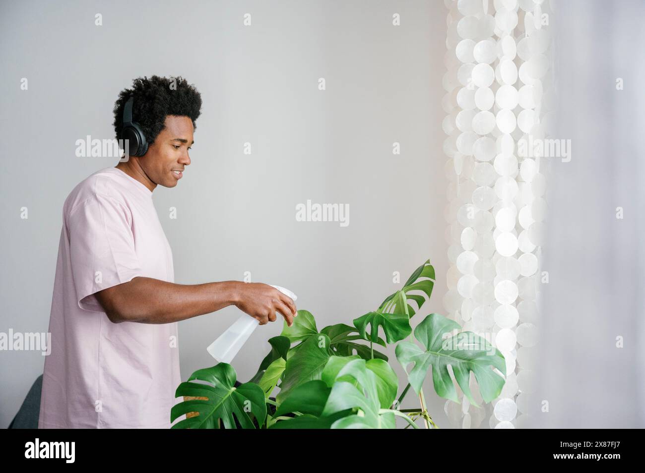Lächelnder Mann, der Musik hört und eine Pflanze mit Sprühflasche zu Hause tränkt Stockfoto