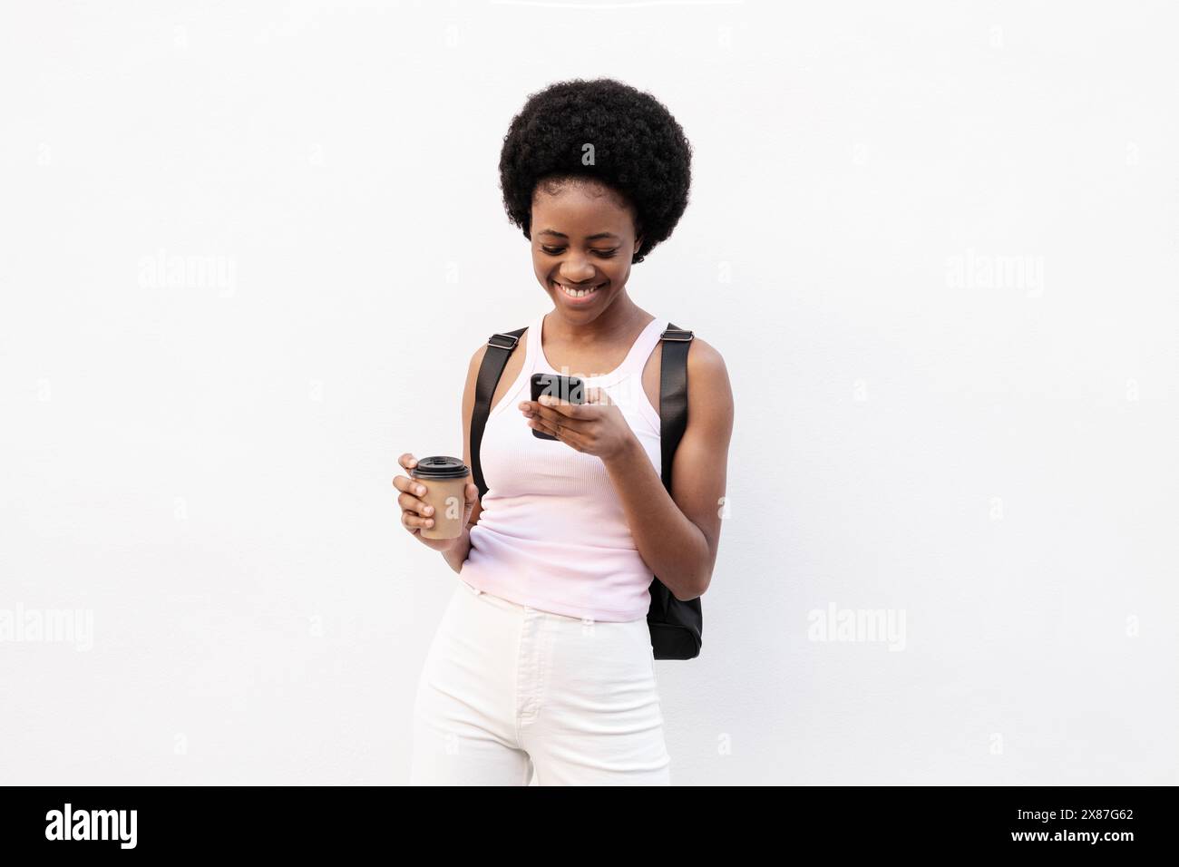 Lächelnde Frau, die Smartphone benutzt und Einwegbecher vor weißem Hintergrund hält Stockfoto