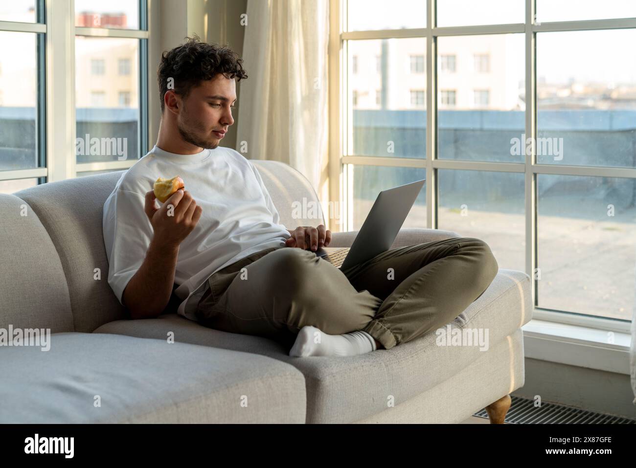 Junger Mann, der einen Laptop benutzt und Apfel zu Hause auf dem Sofa hält Stockfoto