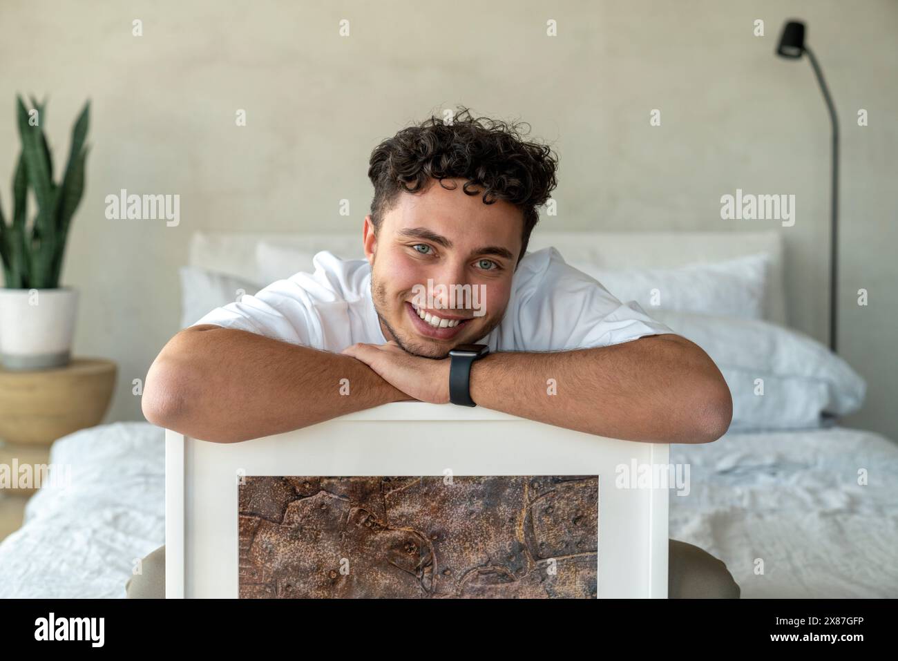 Glücklicher junger Mann, der zu Hause mit Bilderrahmen auf dem Bett sitzt Stockfoto