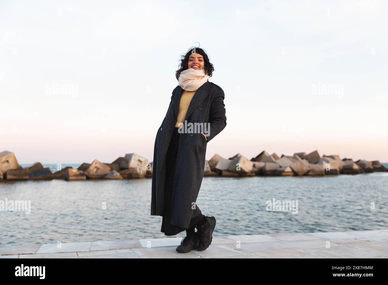 Lächelnde Frau im langen Mantel, die auf der Promenade in der Nähe des Meeres steht Stockfoto