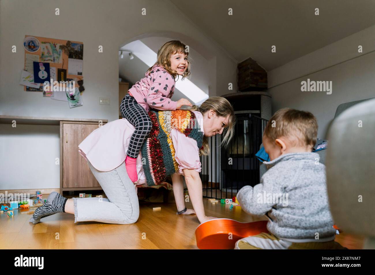 Lächelndes Mädchen reitet auf dem Rücken der Mutter in der Nähe des Jungen, der zu Hause spielt Stockfoto