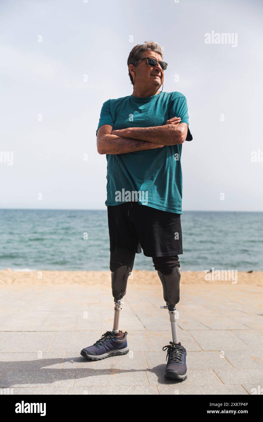 Behinderter Seniorenmann mit prothetischen Beinen, stehend mit gekreuzten Armen am Strand an sonnigen Tagen Stockfoto