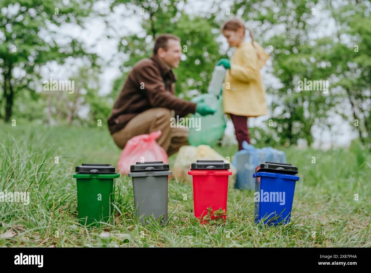 Mehrfarbige Spielzeugmülltonnen mit Vater und Tochter, die Plastikflaschen im Wald sammeln Stockfoto