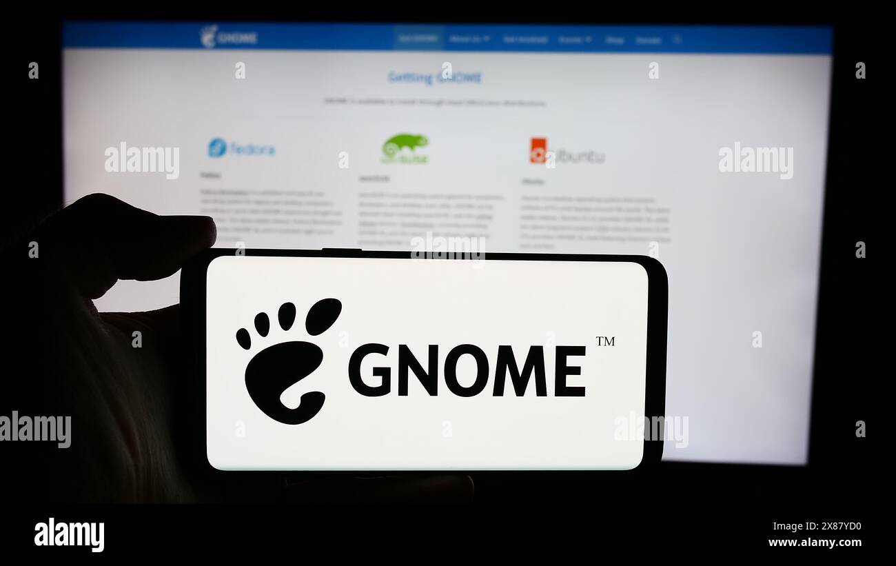 Person, die ein Mobiltelefon mit dem Logo der Open-Source GNU Network Object Model Environment (GNOME) vor der Webseite hält. Schwerpunkt auf dem Display des Telefons. Stockfoto