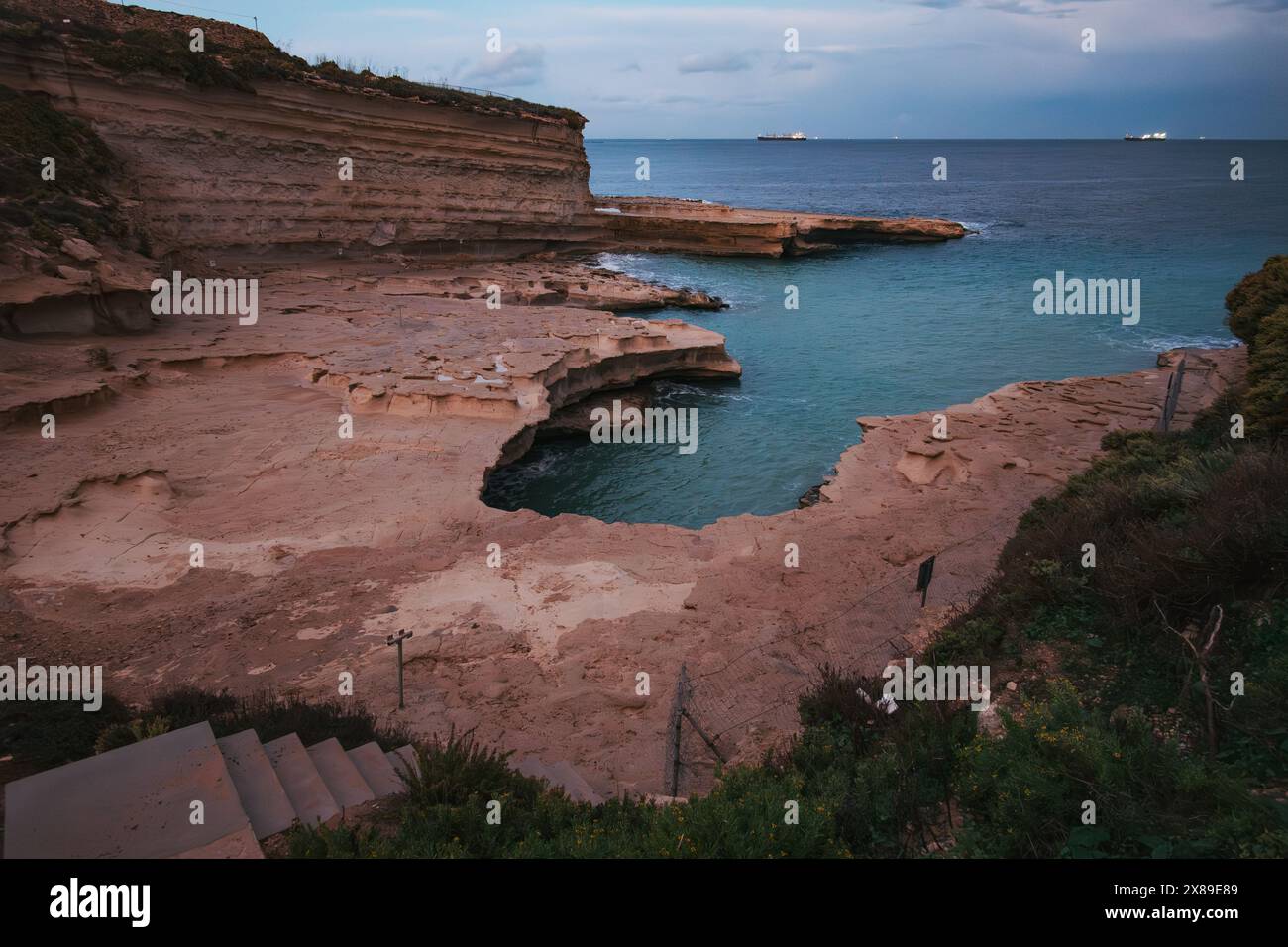 Ruhiger Blick auf Maltas Küstenklippen am St. Peter's Pool, unter dramatischem bewölktem Himmel in der Dämmerung, mit Schiffslichtern in der Ferne Stockfoto