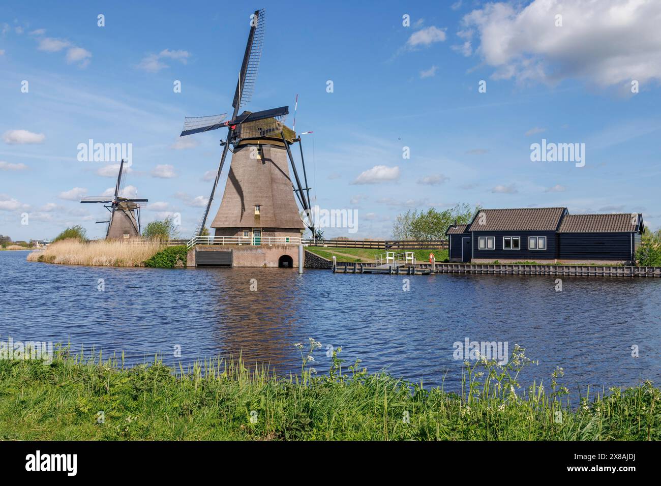 Zwei Windmühlen am Ufer eines Flusses in einer grünen Landschaft mit zusätzlichen Gebäuden, alte Windmühlen an einem Fluss in den Niederlanden mitten in WID Stockfoto